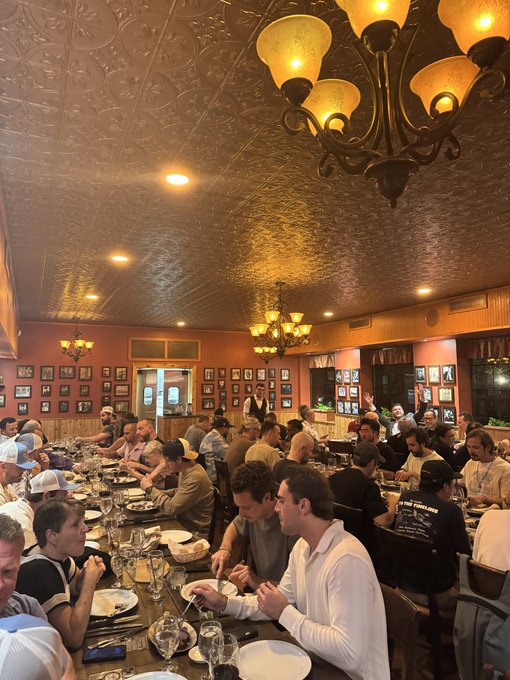 A crowded restaurant with ornate chandeliers, tin ceiling, and framed pictures on the walls is filled with diners at long tables.