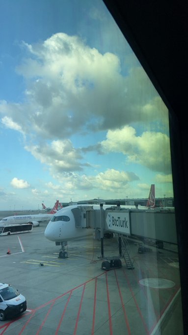 A view through an airport terminal window showing a Turkish Airlines plane (with 'Türkiye'nin... birlikte...' text) parked at a gate with a jet bridge under a cloudy blue sky.