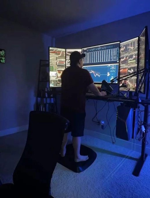 The image shows a man standing at a desk using a computer monitor that displays stock market data. The man appears to be focused on his work or possibly monitoring the stock market, as he has multiple screens in front of him. There are two chairs visible in the room - one near the left side and another closer to the center. A keyboard is also present on the desk next to the computer monitor.