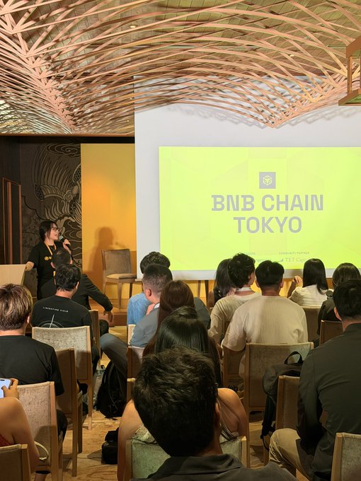 An audience sits facing a presentation screen displaying 'BNB CHAIN TOKYO' in a room with a distinctive woven wood ceiling, while a speaker stands to the left.