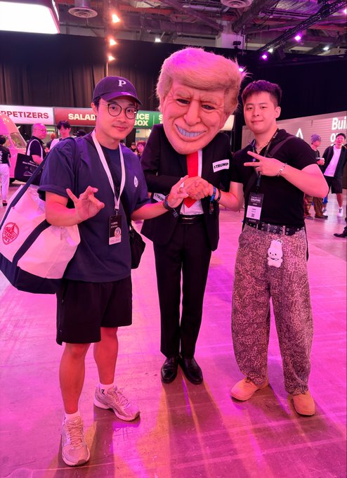 Two young men pose on either side of a person in a suit wearing a large caricature mask of Donald Trump at what appears to be a conference venue.