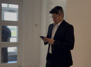 A man in a dark suit stands indoors, looking down at his smartphone with a focused expression.