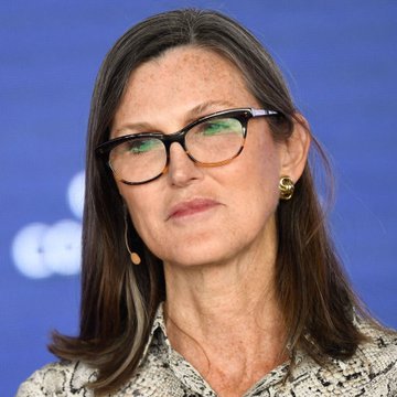 A close-up portrait of a woman with brown hair and black-framed glasses wearing a snakeskin-patterned blouse against a blue background.