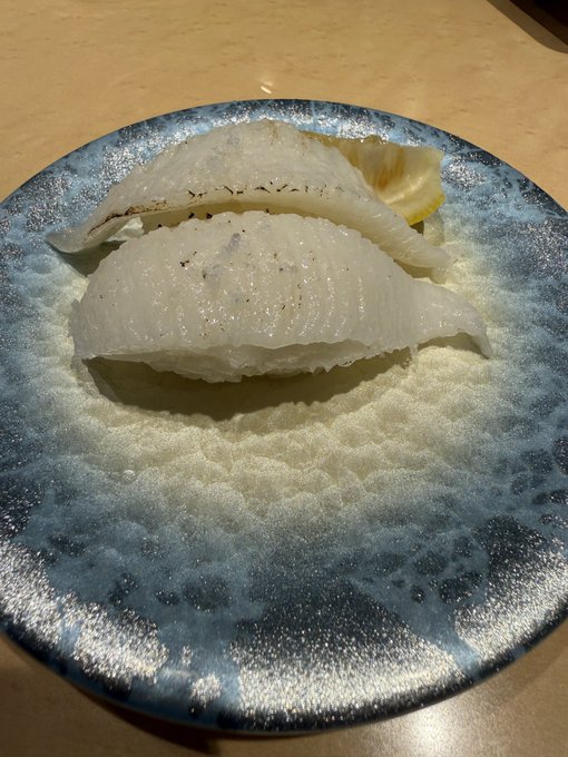 Two pieces of white sushi, likely flounder or halibut nigiri, rest on a textured blue ceramic plate with a piece of pickled ginger.