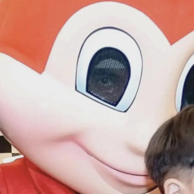 A young person with dark hair partially visible in the lower right corner posing in front of a large cartoon character face with big white eyes and an orange background.