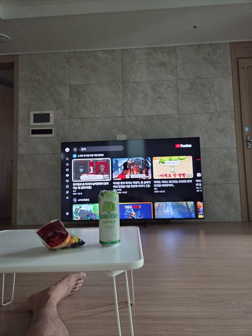 A can of Somersby Apple cider and snacks sit on a small white table in front of a large Samsung TV displaying YouTube Premium in Korean, with a person's bare feet visible at the bottom of the frame. The cozy scene illustrates the lifestyle described in the post—a relaxed evening at home with premium amenities and a drink in hand, fitting the "조건" about having a nice big TV and enjoying a comfortable life.