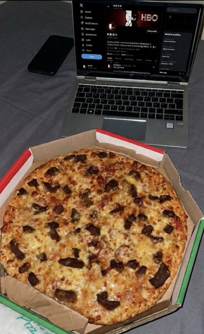 A photograph of a meat pizza in a delivery box placed in front of a laptop displaying the HBO Max interface, with a smartphone lying nearby on a gray surface.