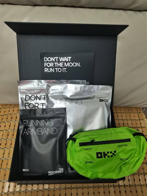 An open black gift box containing OKX-branded running merchandise including a green waist pack ('Don't Run for the Moon, Run to It') and silver packages.