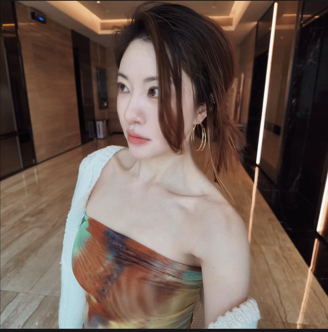 A selfie of a young woman with long brown hair, gold hoop earrings, wearing a colorful tie-dye strapless top and white cardigan, taken in a modern lobby with marble floors.