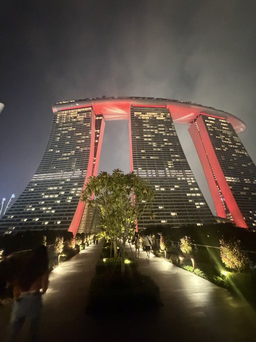 A nighttime photo of the Marina Bay Sands hotel in Singapore illuminated with red lighting, viewed from a landscaped pedestrian walkway.