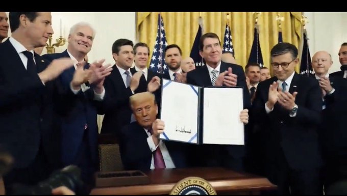 A group of men in suits applauding as Donald Trump, seated at the Resolute Desk, holds up a signed document during a ceremony in the White House.