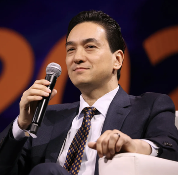 A smiling man in a dark suit and patterned tie holds a microphone while seated, appearing to address an audience during a speaking event. The image likely depicts Simon Gerovich, the CEO of Tokyo-based Metaplanet, whose company recently faced a nearly 10% stock decline amid controversies over his expanded executive option pool and ties to MMXX Ventures.
