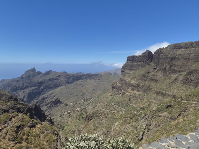 A scenic mountain landscape view from a high vantage point, showing rugged rocky cliffs and peaks with sparse green vegetation under a clear blue sky.