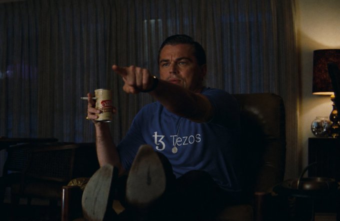 Leonardo DiCaprio as Jordan Belfort in The Wolf of Wall Street, wearing a blue 'Tezos' t-shirt, holding a mug and cigarette while pointing in a dimly lit room.
