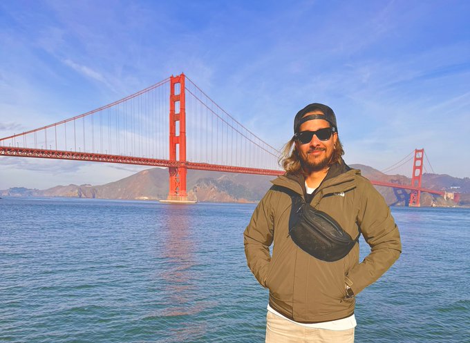 A man wearing a cap, sunglasses, and an olive-green jacket with a black crossbody bag stands in front of the Golden Gate Bridge on a clear, sunny day.