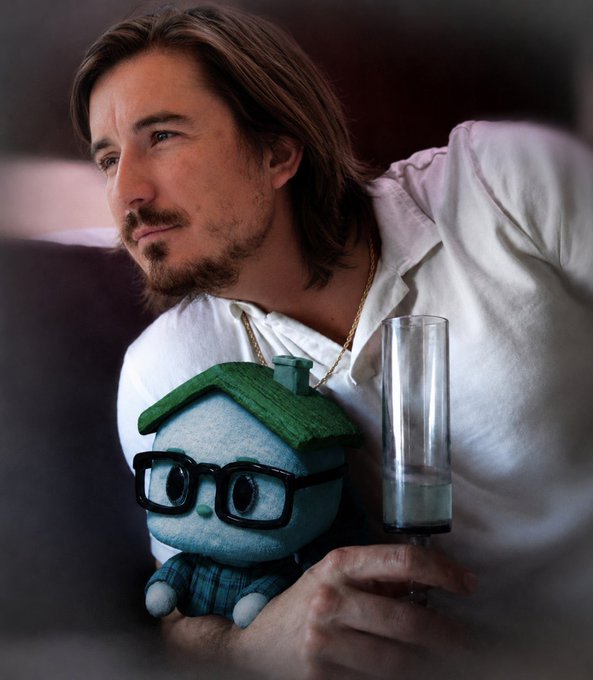A man with long brown hair and a goatee, wearing a white shirt and gold chain, holding a glass of water and a blue plush toy with glasses and a green roof, looking off to the side.