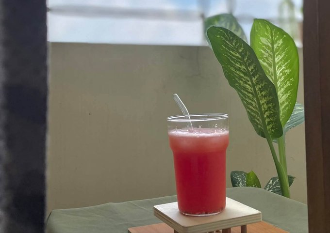 A glass of pink juice with a straw sits on a wooden coaster beside a dieffenbachia houseplant near a window.