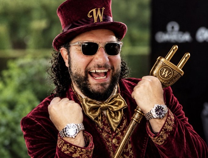 A man in Willy Wonka-style attire, including a maroon velvet top hat with 'W' emblem, sunglasses, and gold bow tie, holds a large gold plug-shaped cane while wearing two luxury watches.