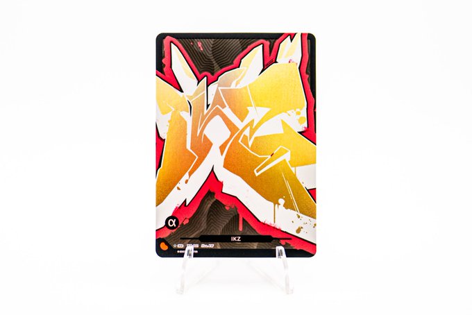 A graffiti-style trading card displayed on a clear stand features yellow and orange abstract street art designs with the text 'α' and 'IKZ' at the bottom.