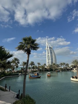 A scenic daytime view of the Burj Al Arab hotel in Dubai seen from across a waterway, framed by palm trees with a small boat on the water.