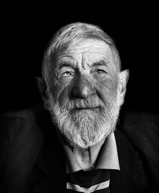 A black-and-white portrait photograph of an elderly man with a white beard and weathered face looking upward against a dark background.