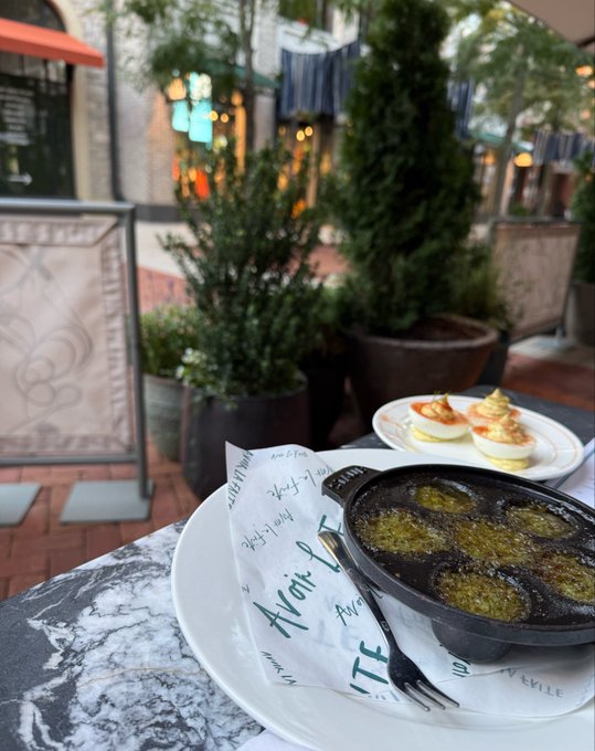 A photograph taken at an outdoor restaurant table shows a cast-iron pan of escargot and a plate of deviled eggs on the Avoca-branded paper liner.