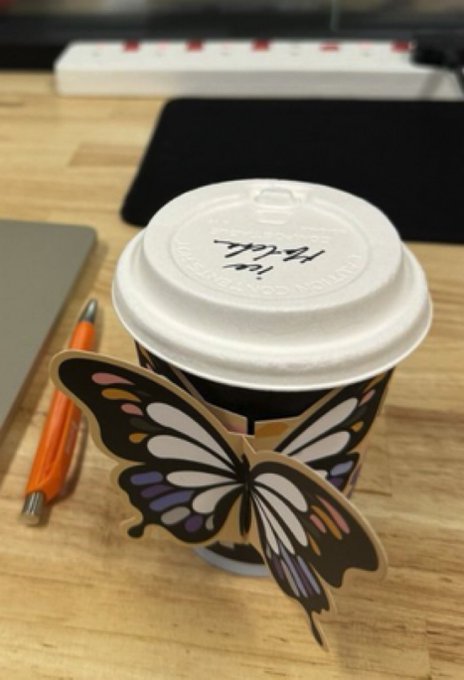 A coffee cup with a butterfly-themed sleeve sits on a wooden desk, with a pen and keyboard visible in the background. The cup lid bears the handwritten text "MERIT/1" alongside what appears to be a logo, possibly referencing Merit Circle or a similar crypto project—highlighting the poster's point that real-world crypto marketing experience matters more than academic credentials.