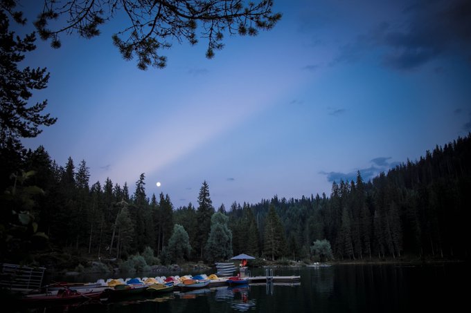 A serene twilight scene of a lake surrounded by dense forest, with colorful kayaks docked at a pier and a moon visible in the dusky sky.