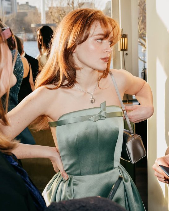 A young woman with flowing auburn hair and bangs gazes to the side in a sage green strapless dress adorned with a small bow, wearing a delicate pendant necklace and carrying a gray handbag, embodying the ethereal beauty described in the caption.