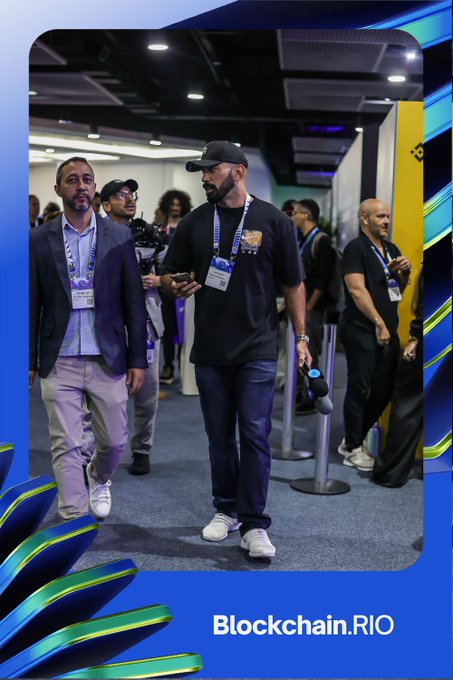 Two men walking through a busy conference venue, framed in a blue-and-green Blockchain.RIO event graphic.