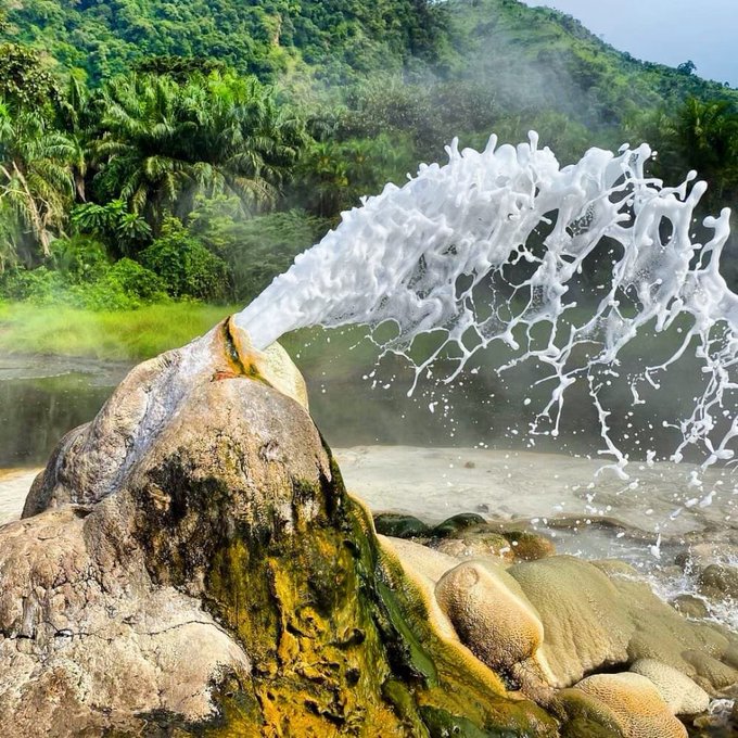 A powerful blast of water or steam erupts from a moss-covered rocky outcrop in a lush tropical landscape, symbolizing the forceful energy of collaborative creation. The image visually represents the post's message about builders on CT (Crypto Twitter) "cooking" together—rising up with intensity and momentum through connection, support, and shared growth.