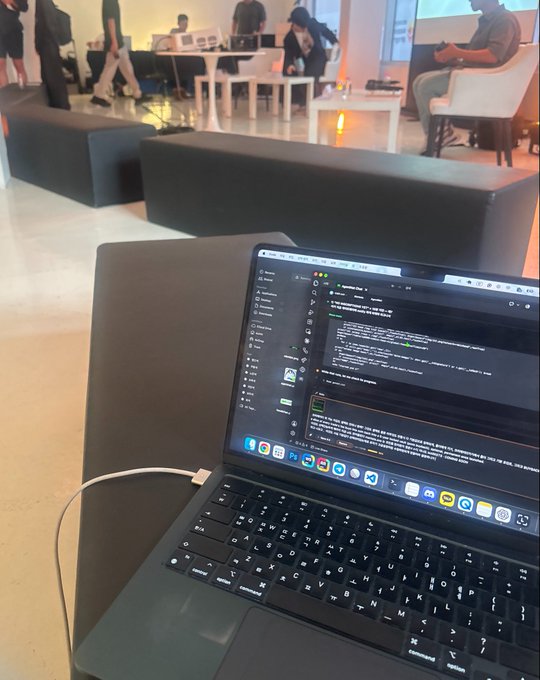 A laptop on a black seat displaying what appears to be a Slack conversation, with a blurred background showing people at what looks like a tech event or office space.