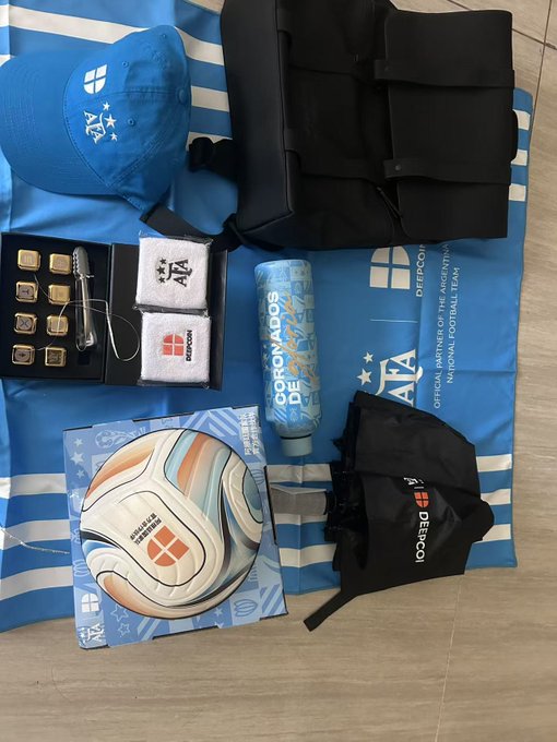 The image shows a collection of blue-themed merchandise items including a cap, backpack, branded chocolates/gold pieces, a towel, a "DeepCoin" branded bottle, a soccer ball featuring the ATA (Asociación del Fútbol Argentino) logo, a DeFiCoin drawstring bag, and an umbrella, all displayed on an AFA Argentina National Football Team banner. This perfectly matches the post describing a care package received from @deepcoin_cn containing branded soccer merchandise, custom water bottle, crypto gold ice stones, umbrella, and wristbands.