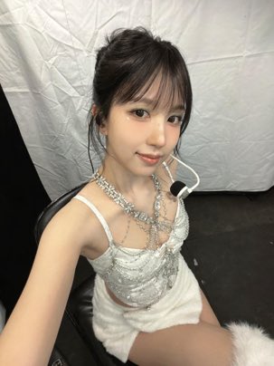 A young woman in a sparkly silver and white outfit with elaborate rhinestone necklaces poses for a portrait against a white backdrop.