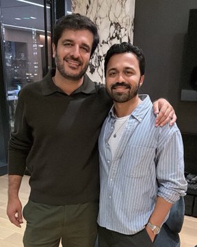 Two smiling men pose together indoors with their arms around each other; the man on the left wears a dark sweater and the man on the right wears a striped button-down shirt.