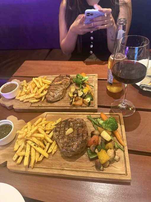 A steak dinner with fries and vegetables is served on a wooden board alongside a glass of red wine and a bottle, while a person uses their phone at the table. The caption combines the luxurious meal imagery with trading results for $BTC and $ETH, suggesting the celebratory dinner coincides with a profitable start to the trading week.