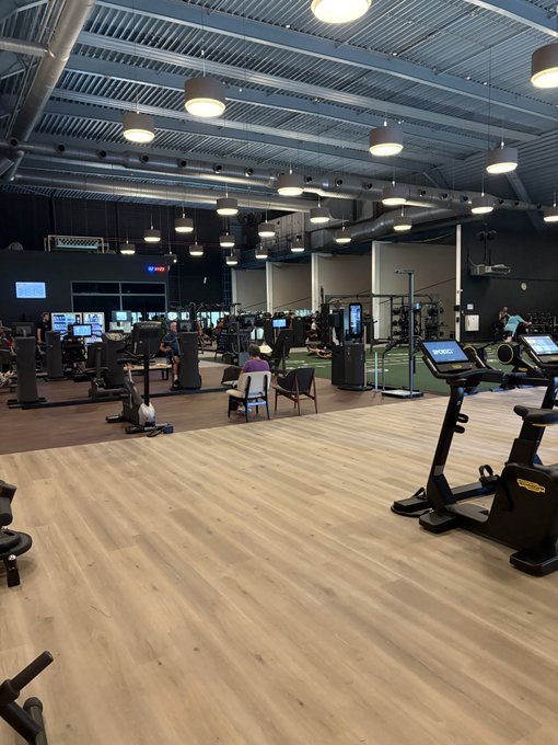 A spacious, modern gym interior with industrial-style ceiling, pendant lighting, and various cardio equipment including treadmills and stationary bikes. Several people are using the machines, with rows of equipment visible in the background.