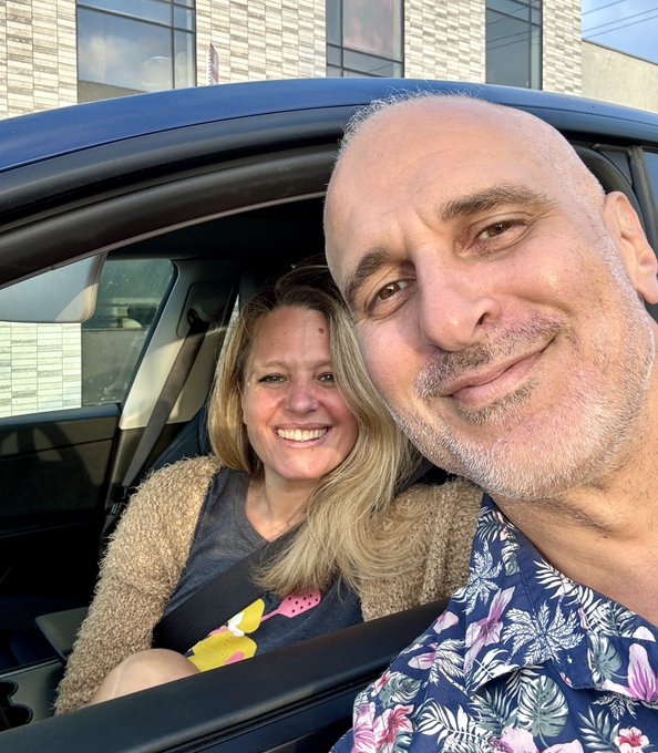A selfie of a smiling couple inside a car, with the man in a blue floral shirt in the foreground and the woman in a tan cardigan smiling behind him.