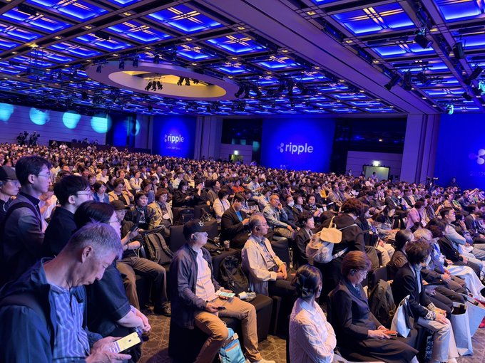 A large audience sits in a dimly lit, blue-hued conference hall with "ripple" logos displayed prominently on the stage backdrop.