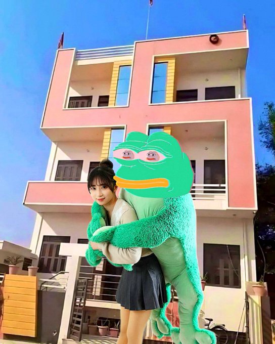 A young woman in a school uniform pose is embraced by a person in a green Pepe the Frog costume in front of a pink building.