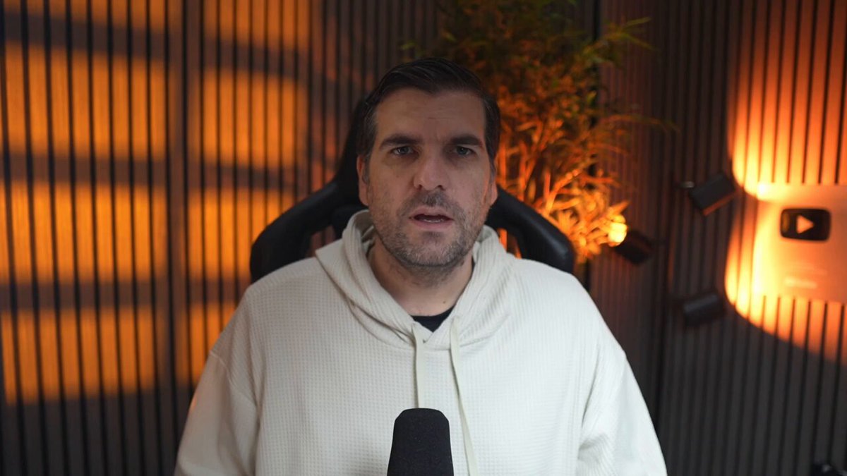 The image shows an adult male wearing a white hoodie, sitting in front of a microphone with a laptop behind him. The man appears to be talking or giving a speech, possibly at a live event or online. The background features a wall with orange lights that create a warm and inviting atmosphere for the audience. The post includes information about Bitcoin and XRP, suggesting an interest in cryptocurrency markets and their potential impact on the economy.