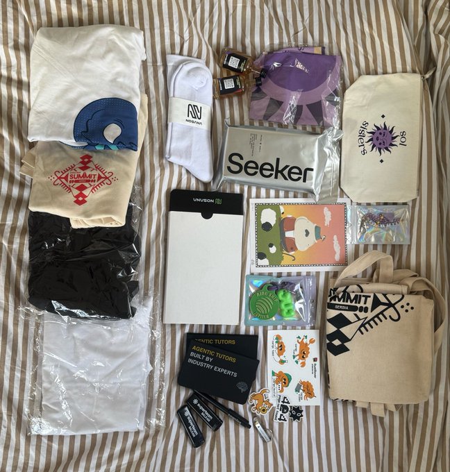 A flat-lay collection of merchandise items including t-shirts, socks, stickers, a tote bag, and badges spread out on a striped fabric surface, with visible text like 'Seeker', 'SUMMIT', and 'ARCTIC TUTORS'.
