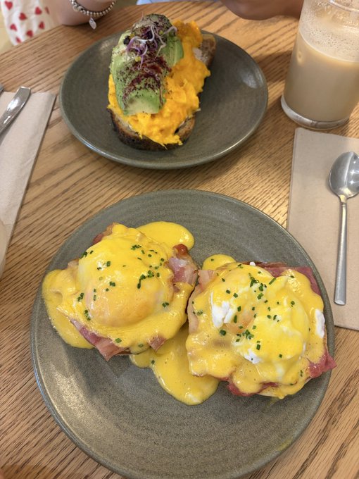 A breakfast spread on a wooden table featuring eggs Benedict with ham and hollandaise sauce on a green plate, alongside a slice of avocado toast topped with scrambled eggs and microgreens on another green plate, plus a glass of latte.
