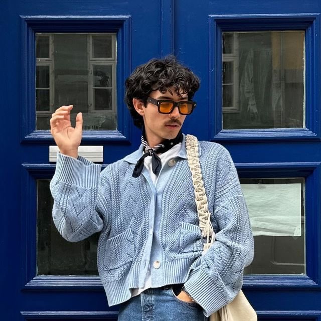 A stylish young man with curly hair, sunglasses, and a black neck scarf poses in a light blue cable-knit cardigan in front of a vibrant blue paneled door.