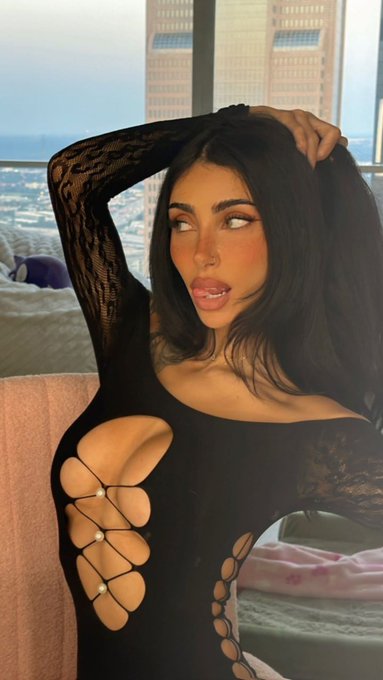 A young woman with long dark hair takes a selfie in a black long-sleeve top with large cutouts, set against a window with a city skyline view in the background.