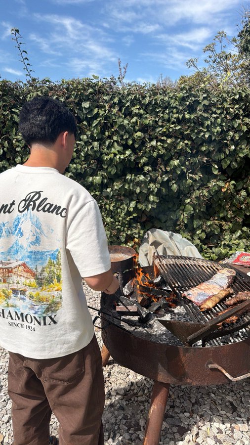 A young man wearing a 'Mont Blanc CHAMONIX SINCE 1924' t-shirt tends to meat cooking on a charcoal grill outdoors.