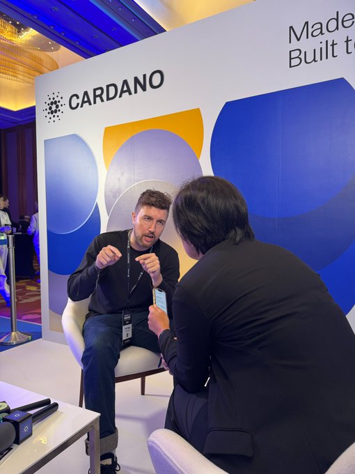 Two men converse in chairs in front of a 'CARDANO' branded backdrop at an event, with text reading 'Made' and 'Built to'.