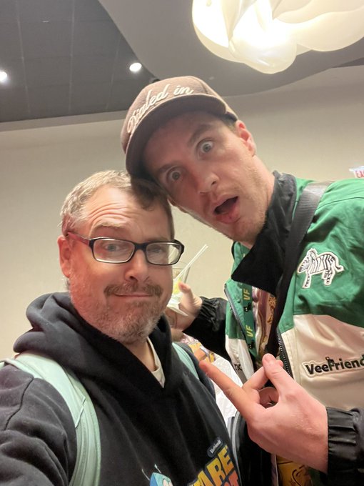 A selfie of two men at what appears to be a convention; one wears a 'VeeFriends' jacket and a 'finished in' cap, while the other makes a peace sign in a black hoodie.