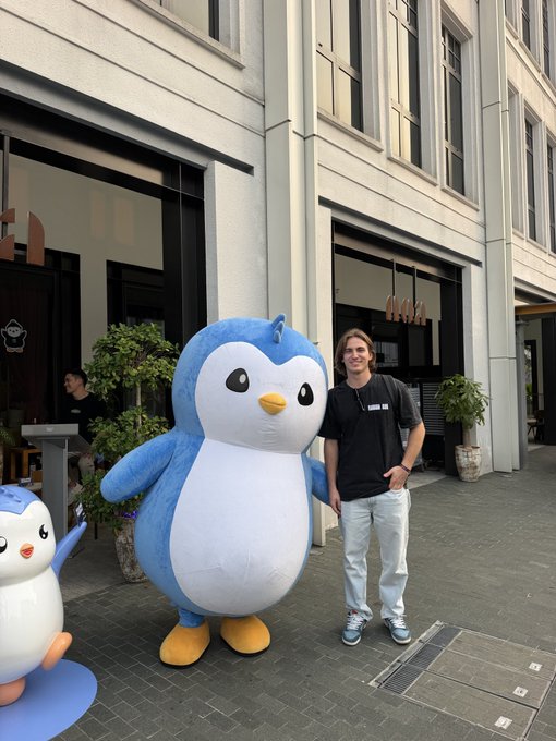 A young man in a black t-shirt and light pants stands smiling next to a large blue and white penguin mascot (resembling Pingu) on a sidewalk outside a building, with a smaller penguin figure nearby.