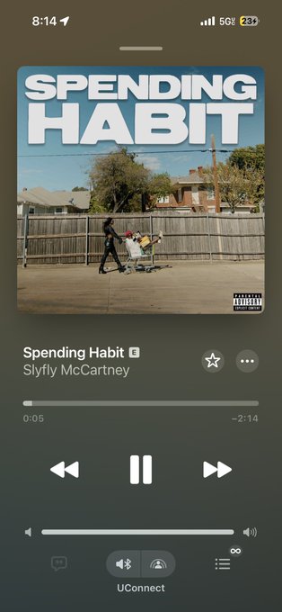 A phone screenshot of Apple Music playing the song 'Spending Habit' by Slyfly McCartney, showing the album cover of a person pushing a shopping cart.